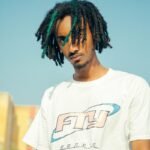 Close-up portrait of a young man with dreadlocks wearing a casual white shirt in daylight.
