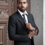 Elegant man in a black suit posing confidently outdoors with a serious expression.